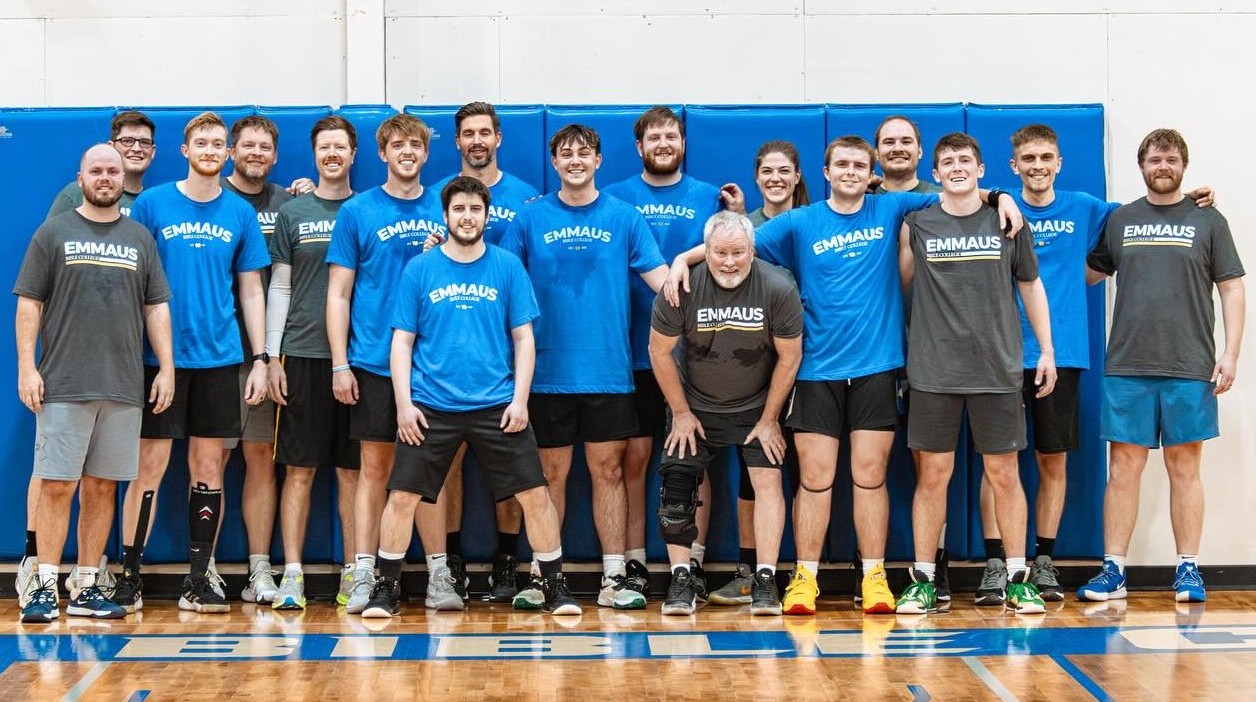 A group of men of varying age in Emmaus t-shirts pose against a gym wall