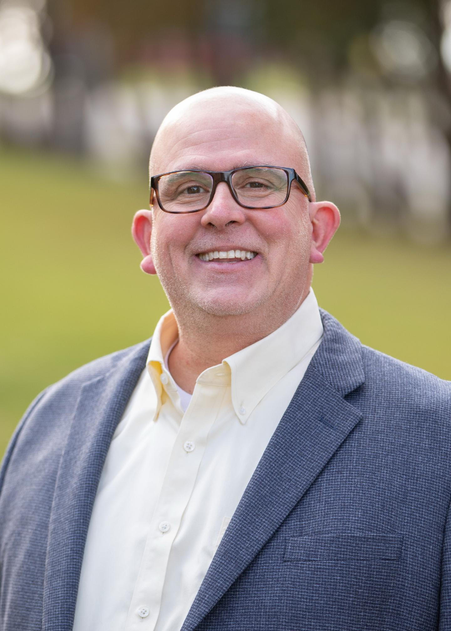 A smiling bald man with glasses wearing a blue suit jacket and white shirt