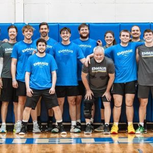 Men in blue and gray Emmaus shirts stand along the wall in the gym
