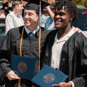 Two young men stand in graduation caps and gowns hold up their diplomas