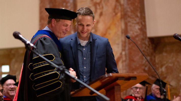 President in graduation regalia shakes hand of man receiving award
